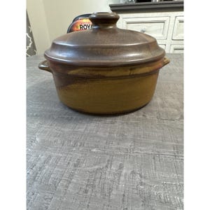 Pode incluir: Uma caçarola de cerâmica marrom com tampa. O prato tem formato redondo com duas alças e uma tampa com botão. O exterior tem um esquema de cores salpicadas e terrosas. O prato está sobre uma superfície cinza.