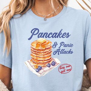 Könnte beinhalten: Hellblaues T-Shirt mit dem Text "Pancakes & Panic Attacks" und einer Grafik von Pfannkuchen mit Butter, Sirup und Blaubeeren. Das Shirt hat auch den Text "buy one get one".