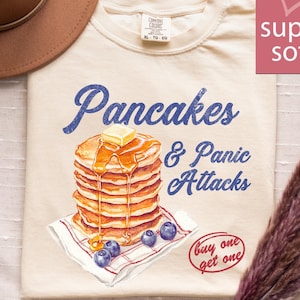May include: Cream-colored t-shirt with the text "Pancakes & Panic Attacks" in blue script. A stack of pancakes with butter and syrup is pictured, along with blueberries. The shirt also has the text "buy one get one".