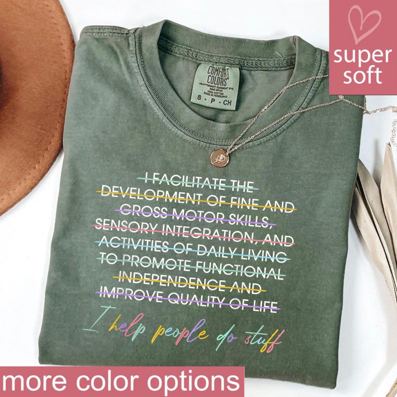May include: Olive green Comfort Colors t-shirt with the text "I FACILITATE THE DEVELOPMENT OF FINE AND GROSS MOTOR SKILLS...I help people do stuff." A gold necklace and a pink heart graphic are also visible.