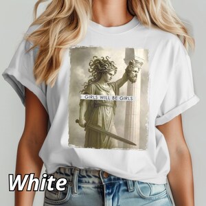 May include: White t-shirt featuring a graphic of a classical statue holding a sword and a severed head. The text "GIRLS WILL BE GIRLS" is printed on the graphic. The word "White" is printed at the bottom of the shirt.