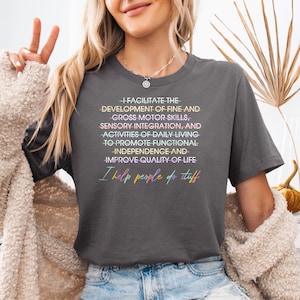May include: Dark gray t-shirt with the text "I FACILITATE THE DEVELOPMENT OF FINE AND GROSS MOTOR SKILLS... I help people do stuff" in colorful text. The shirt is a crew neck style.