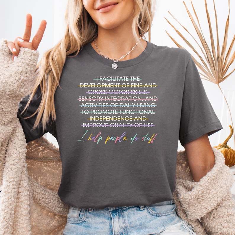 May include: Dark gray t-shirt with the text "I FACILITATE THE DEVELOPMENT OF FINE AND GROSS MOTOR SKILLS... I help people do stuff" in colorful text. The shirt is a crew neck style.