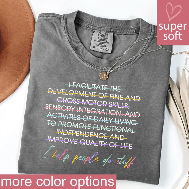 May include: A gray Comfort Colors t-shirt with the text "I FACILITATE THE DEVELOPMENT OF FINE AND GROSS MOTOR SKILLS...I help people do stuff" in colorful lettering. A silver necklace with a pendant rests on the shirt.