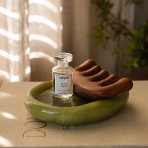 May include: A small clear glass perfume bottle labelled REPLICA Beach Walk Maison Margiela, placed next to a curved wooden hair comb on a smooth, round, olive green ceramic tray resting on a beige book titled DONATELLO SCULPTURES.