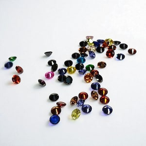 May include: A scattering of colorful gemstones in various shapes and sizes. The gems are in shades of red, blue, green, yellow, and black, with a few pink and brown stones. The gemstones are scattered on a white surface.