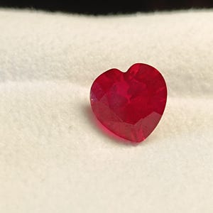 May include: A heart-shaped, ruby-red gemstone with multiple facets. The gem is set against a white, textured background. The heart shape is clearly defined, with the facets catching the light.