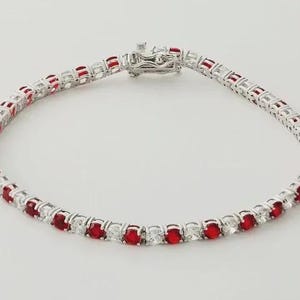 Könnte beinhalten: Ein silberfarbenes Tennisarmband mit einem sich wiederholenden Muster aus runden, leuchtend roten Steinen, die sich mit klaren, funkelnden Steinen abwechseln. Das Armband wird durch einen metallischen Kastenschloss-Verschluss geschlossen.