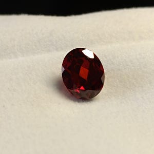 May include: A faceted, oval-shaped, deep red gemstone. The gem is set against a white, textured background. The facets catch the light, creating a brilliant display of color and sparkle. This gemstone could be used in jewelry.