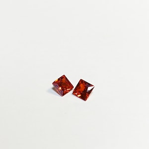 May include: Two square-cut, vibrant orange gemstones with faceted surfaces. The gems are displayed against a white background, showcasing their colour and clarity. Ideal for jewellery making or collecting.
