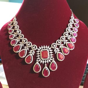 May include: A dazzling necklace featuring a central rectangular ruby surrounded by sparkling white stones. The necklace has a symmetrical design with teardrop-shaped rubies and circular accents, displayed on a red velvet surface.