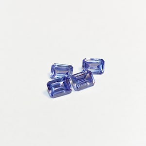 May include: Four rectangular, faceted, blue gemstones are arranged on a white surface. The gems have a slight violet hue and are cut with clean lines, reflecting light. The gems are likely for jewelry making or collecting.