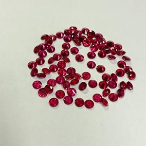 May include: A collection of small, round, faceted rubies. The gemstones are a deep, vibrant red color and are scattered across a white surface. The rubies vary slightly in size and shape, with a polished, reflective surface.