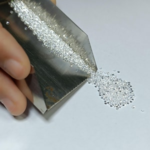 May include: Close-up of a metal scoop filled with numerous small, clear diamonds. The diamonds are spilling out onto a white surface. The image highlights the brilliance and clarity of the gemstones.