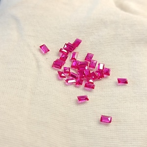 May include: A collection of small, rectangular, bright pink gemstones scattered on a white cloth. The gemstones have a polished, reflective surface and are of varying sizes. The cloth has a slightly textured appearance.