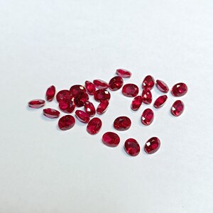 May include: A collection of oval-cut, deep red gemstones scattered on a white surface. The gems vary slightly in size and shape, with a polished, reflective surface. The overall effect is a display of vibrant, natural rubies.