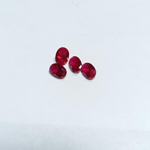 Puede incluir: Cinco rubíes ovalados y facetados. Las piedras preciosas son de un color rojo intenso y vibrante y se muestran sobre un fondo blanco. Los rubíes están dispuestos en un patrón ligeramente disperso.