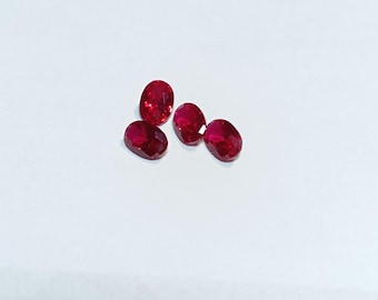 May include: Five oval-cut, faceted rubies. The gemstones are a deep, vibrant red color and are displayed against a white background. The rubies are arranged in a slightly scattered pattern.