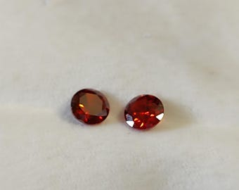 Pode incluir: Duas pedras preciosas ovais, vermelho profundo. As gemas são facetadas, captando a luz e exibindo um brilho brilhante. Elas são colocadas sobre um fundo branco macio, destacando sua cor rica e clareza. Estas pedras preciosas são adequadas para a fabricação de joias.