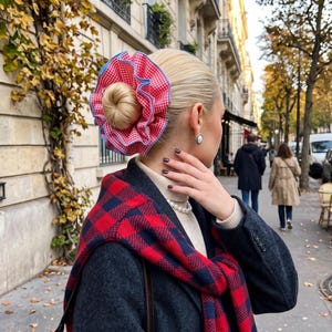 Peut inclure: Un grand chouchou à volants avec un motif vichy rouge et blanc et un bord bleu. Le chouchou est porté dans un chignon haut, associé à un pull à col roulé crème, un manteau gris foncé et une écharpe à carreaux rouge et bleu marine.