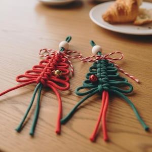 Könnte beinhalten: Zwei handgefertigte Makramee-Weihnachtsbaumschmuck. Einer ist rot, der andere grün, jeweils mit einer kleinen goldenen Glocke und einer Holzperle. Der Schmuck liegt auf einem Holztisch, im Hintergrund Kaffeetassen und ein Croissant.