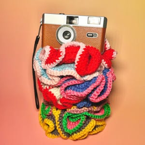 Peut inclure: Un appareil photo vintage marron et argenté avec une sangle noire repose sur une pile de crochet coloré fait à la main. Le crochet présente des fils rouges, bleus, roses, jaunes et verts, créant un affichage vibrant et texturé. L'appareil photo a un objectif sans mise au point.