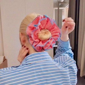 May include: A red and white gingham scrunchie with blue trim, styled in a bun. The scrunchie is the main feature, with a light-coloured bun at the centre. The person is wearing a blue and white striped shirt.
