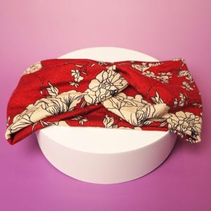 Op de afbeelding: Een rode bloemenhoofdband met een gedraaid knoopontwerp. De hoofdband heeft een cr&egrave;mekleurig bloemenpatroon op een rode achtergrond. Het accessoire wordt gepresenteerd op een witte cilindrische standaard tegen een paarse achtergrond.