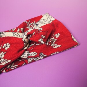 Op de afbeelding: Rode en cr&egrave;mekleurige haarband met bloemenprint. De haarband heeft een gedraaid knoopontwerp en is gemaakt van een zachte, bedrukte stof. Het bloemenpatroon bevat witte bloemen en bladeren op een rode achtergrond. De haarband wordt getoond tegen een paarse achtergrond.