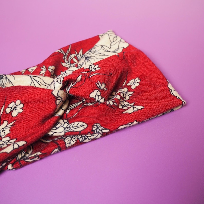 Op de afbeelding: Rode en cr&egrave;mekleurige haarband met bloemenprint. De haarband heeft een gedraaid knoopontwerp en is gemaakt van een zachte, bedrukte stof. Het bloemenpatroon bevat witte bloemen en bladeren op een rode achtergrond. De haarband wordt getoond tegen een paarse achtergrond.
