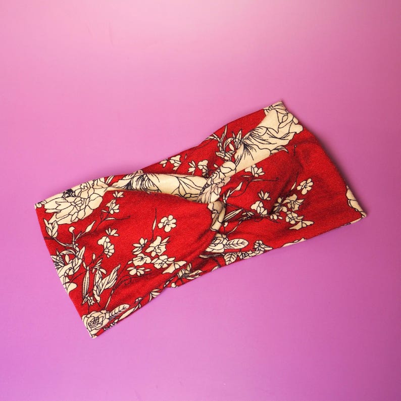 Op de afbeelding: Rode hoofdband met cr&egrave;mekleurige bloemenprint. De hoofdband heeft een gedraaid knoopontwerp en is gemaakt van een zachte stof. Het bloemenpatroon bevat witte bloemen en groene bladeren op een rode achtergrond. Geschikt voor kapsels.