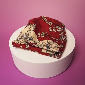 Op de afbeelding: Een opgevouwen rode beanie met een bloemenpatroon in cr&egrave;me en zwart. De hoed is gemaakt van een zachte stof en wordt gepresenteerd op een witte cilindrische sokkel tegen een roze en paarse achtergrond.