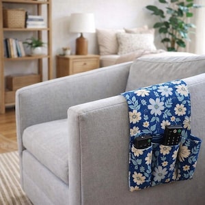 Puede incluir: Un organizador de control remoto con estampado floral azul, colocado sobre el brazo de un sillón gris tapizado. El organizador tiene tres bolsillos, cada uno con un control remoto. El estampado floral incluye flores blancas y amarillas con hojas verdes.