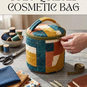 May include: A cylindrical quilted cosmetic bag featuring a geometric patchwork pattern in shades of teal, mustard yellow, and terracotta. The bag has a top handle, a green zipper closure, and is constructed with a grid-like stitched design.