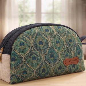 May include: A semi-circular zippered pouch featuring a green and blue peacock feather pattern. The pouch has a dark denim top, a light beige side panel, and a brown leather-like tag on the front with the word Handmade embossed in black text.