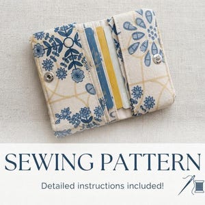 May include: A fabric card holder featuring a cream background with a blue floral pattern and thin yellow lines. The wallet is open, revealing two internal slots containing a blue card and a yellow card, secured by a silver metal snap button closure.