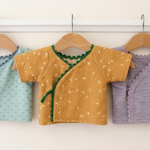 May include: Three short-sleeved wrap shirts hanging on wooden hangers. The left shirt is light blue with a small pattern and dark blue scalloped trim. The middle shirt is mustard yellow with a floral pattern and green trim. The right is purple.