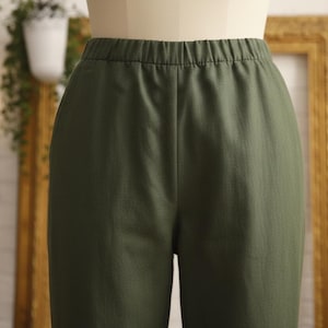 May include: Close-up of olive green trousers with an elasticated waistband. The trousers are made of a smooth fabric and appear to be well-sewn. The trousers are displayed on a mannequin, suggesting they are for sale.