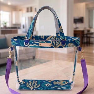 May include: A clear plastic tote bag featuring a blue and teal aquatic pattern with fish and seaweed designs. The bag includes purple fabric straps, gold-toned metal hardware, and a rectangular tan leather-like patch on the front top edge.