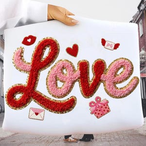 May include: White pillow with the word "Love" in red and pink textured fabric, outlined in gold glitter. The pillow is decorated with embroidered hearts, a kiss, a gift, and an envelope with wings.