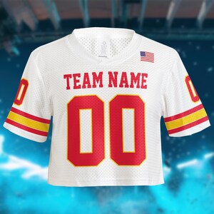 May include: White cropped football jersey with red and yellow accents. Features the text "TEAM NAME" above the number "00" in red. An American flag is on the upper right. The jersey has a v-neck and short sleeves.