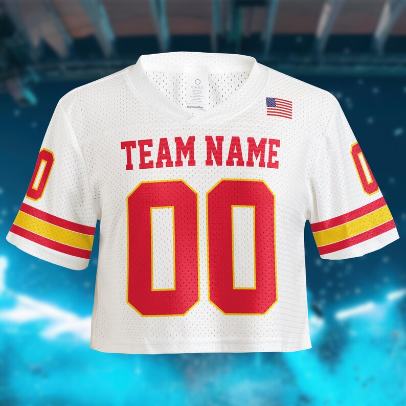 May include: White cropped football jersey with red and yellow accents. Features the text "TEAM NAME" above the number "00" in red. An American flag is on the upper right. The jersey has a v-neck and short sleeves.