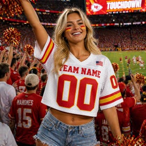 May include: A white football jersey with red and yellow accents, featuring the number "00" and the text "TEAM NAME". The jersey is worn with denim shorts. The background shows a stadium filled with fans.