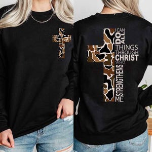 May include: Black long-sleeve sweatshirt featuring a cross design with a camouflage pattern in shades of brown, tan, and white. The back of the shirt displays a larger cross with the text "I CAN DO ALL THINGS THROUGH CHRIST WHO STRENGTHENS ME."