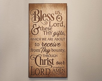 May include: A wooden sign with a brown background and a dark brown inscription. The inscription reads "Bless us, & these Thy gifts, which we are about to receive from Thy bounty, through Christ our LORD. AMEN."