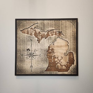 May include: A wooden wall art piece featuring a silhouette of the state of Michigan with a compass and a deer in the forest. The wood has a distressed, rustic look.