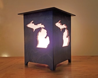 Op de afbeelding: Een houten lamp in de vorm van de staat Michigan met een uitsnijding van de omtrek van de staat. De lamp is aan en geeft een warm licht.