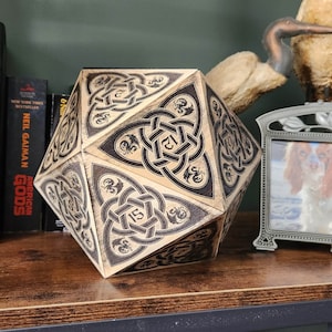 May include: A wooden, multi-sided dice with black and white Celtic knot designs and numbers. The dice is sitting on a wooden shelf with books and a picture frame.