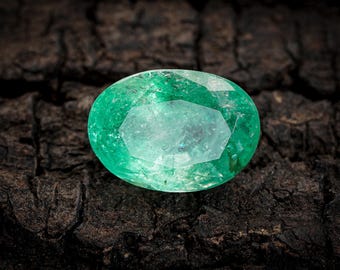 Pode incluir: Uma gema de esmeralda oval, exibindo um tom verde vibrante com clareza interna. A gema é apresentada contra um fundo escuro e texturizado, realçando seu brilho. Uma visão de perto enfatiza as facetas e a cor.