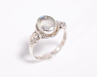 May include: A silver ring with an oval, iridescent gemstone. The ring has an ornate design around the gemstone and on the shoulders of the band. The gemstone has a light blue and white colour.